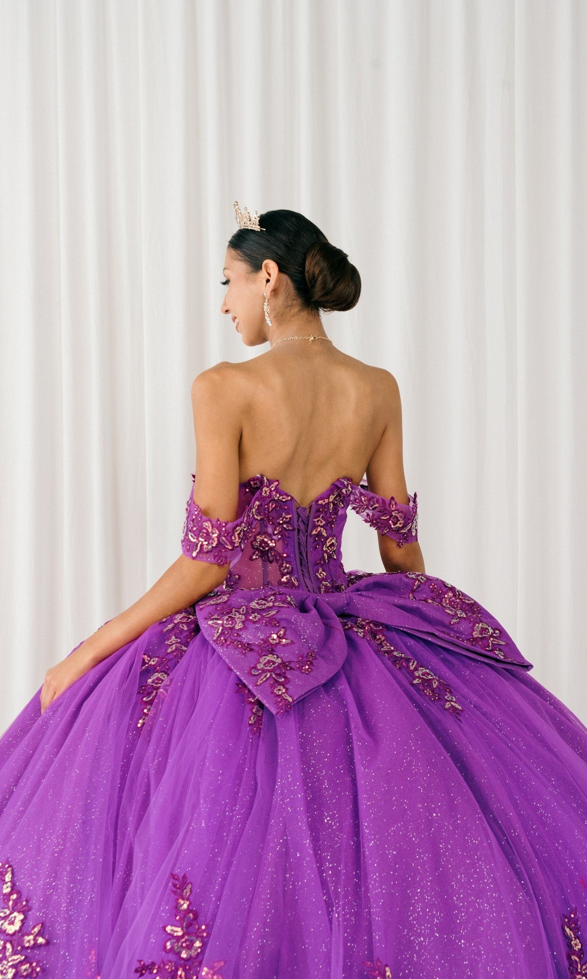 A woman in the purple, off-the-shoulder Quinceanera Dress 8405JQ by Cinderella Couture, featuring a large bow and delicate embellishments, with a tiara, stands facing away before a white curtain backdrop.